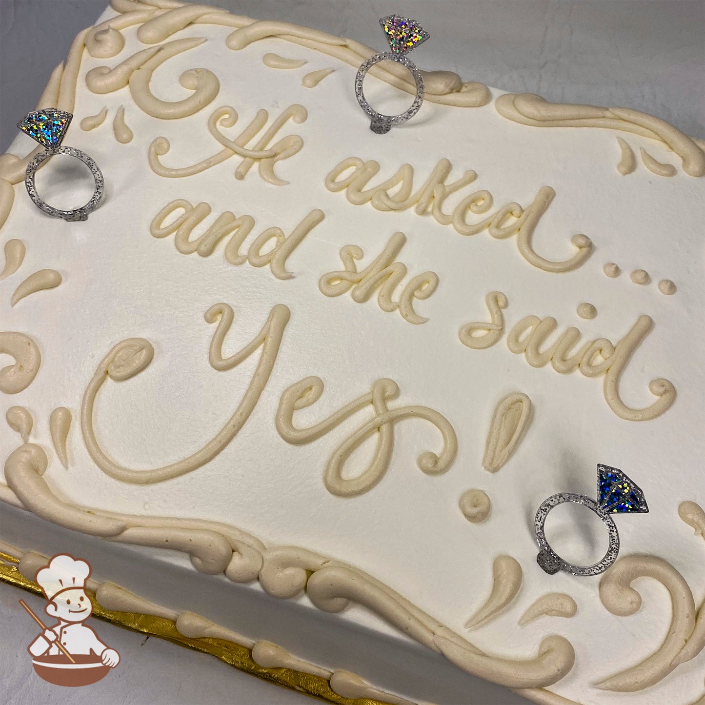 A white sheet cake with cream-colored buttercream scrollwork and borders, decorated with three plastic toy engagement rings featuring glittery gem designs.