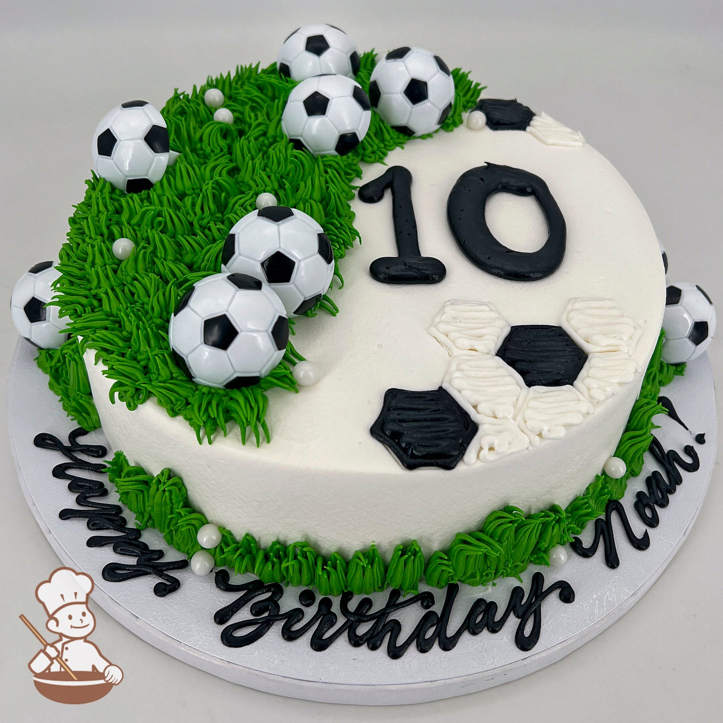 Round cake with white icing, green grass-like piping, plastic soccer ball decorations, black and white hexagon soccer patterns, and the number "10" in black icing.