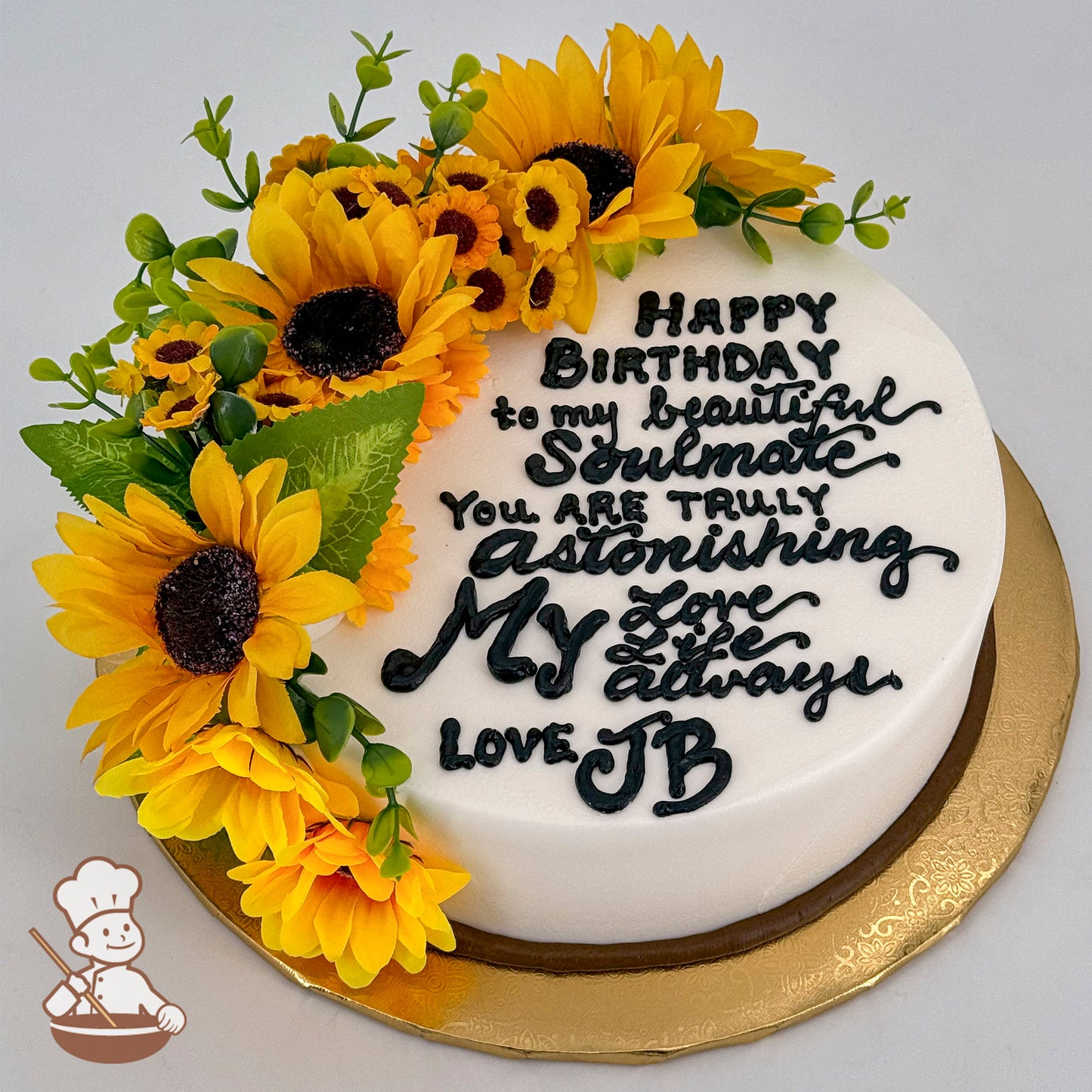 Round white cake with cascading silk sunflowers, including large yellow sunflowers, smaller sunflowers, green leaves, and vines, on a gold base.