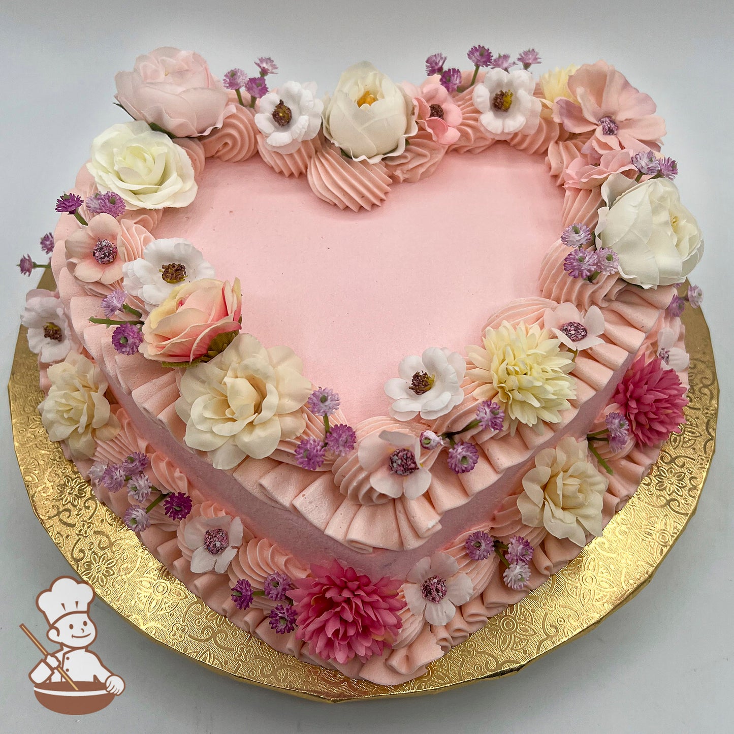 Heart-shaped cake with pink icing, adorned with silk flowers including roses, peonies, and daisies, on a gold-patterned board.