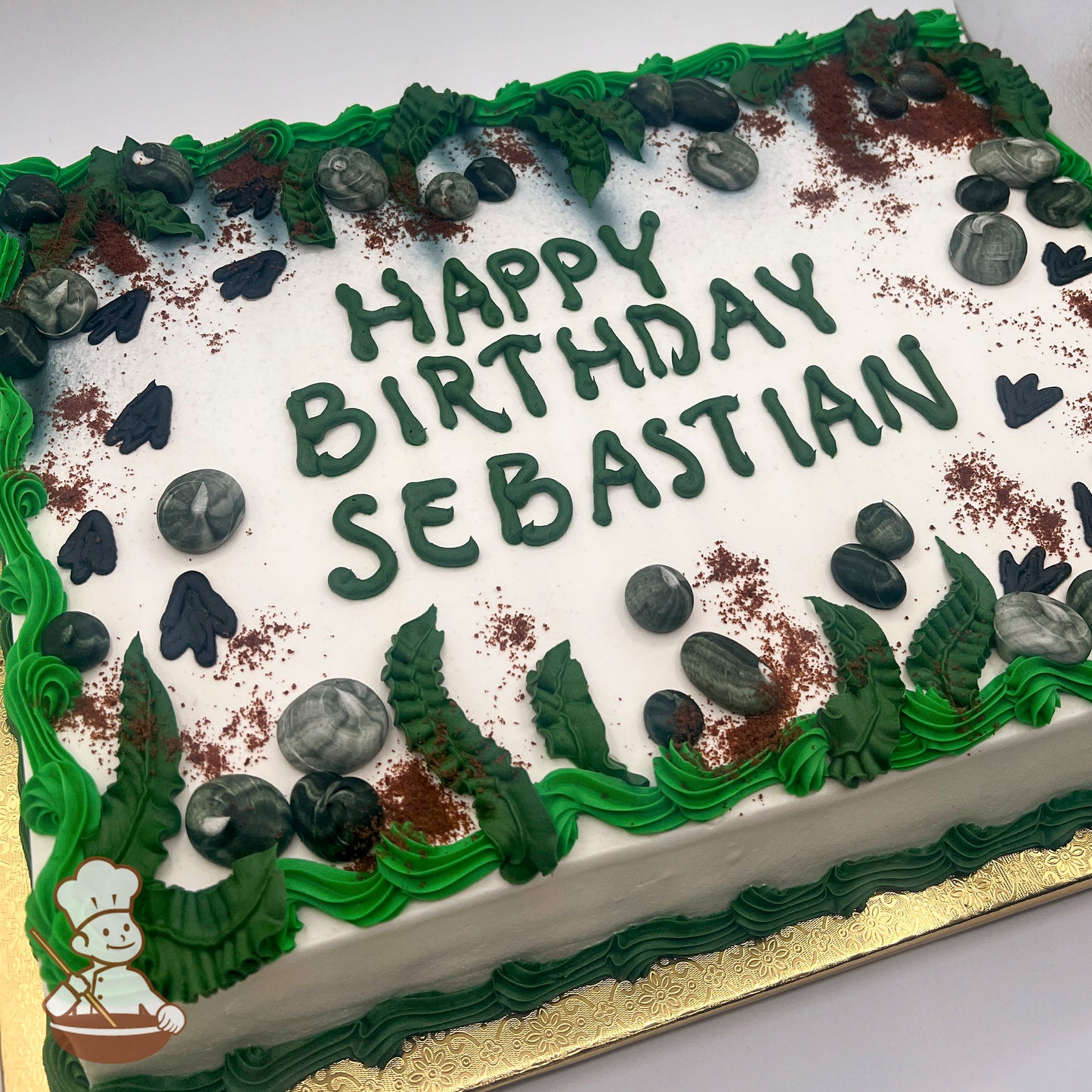 Sheet cake with green buttercream fern leaves, marbled buttercream rocks, cake crumb dirt, and black dinosaur footprints on white frosting.