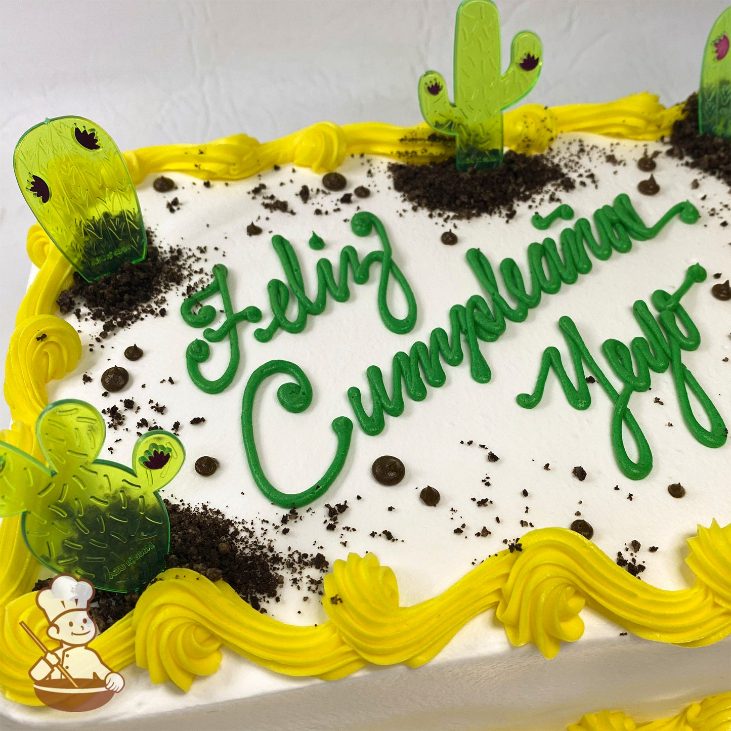 Sheet cake with yellow buttercream border, plastic cactus picks, and cake crumb "dirt" patches. Green writing on top.