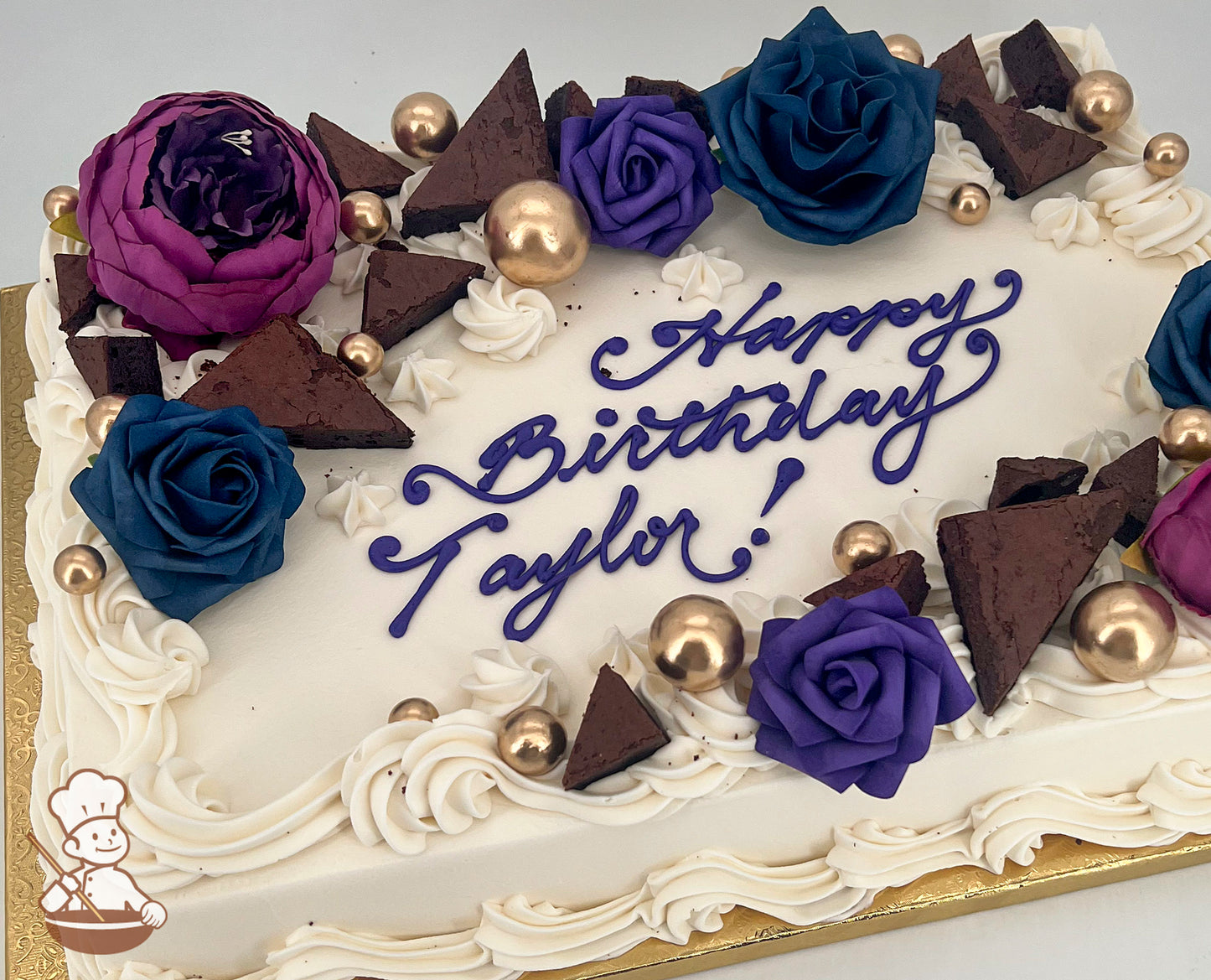 Sheet cake with white frosting, decorated with purple, blue, and magenta silk and foam flowers, chocolate brownie triangles, and gold bubble toppers.