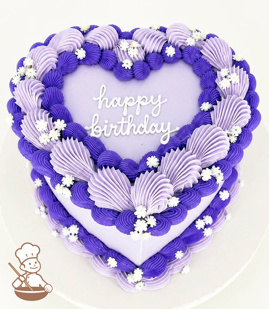 A heart-shaped cake with purple and lavender frosting swirls, white flower decorations, and "happy birthday" written in white icing on top.