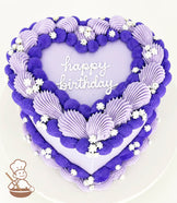 A heart-shaped cake with purple and lavender frosting swirls, white flower decorations, and "happy birthday" written in white icing on top.
