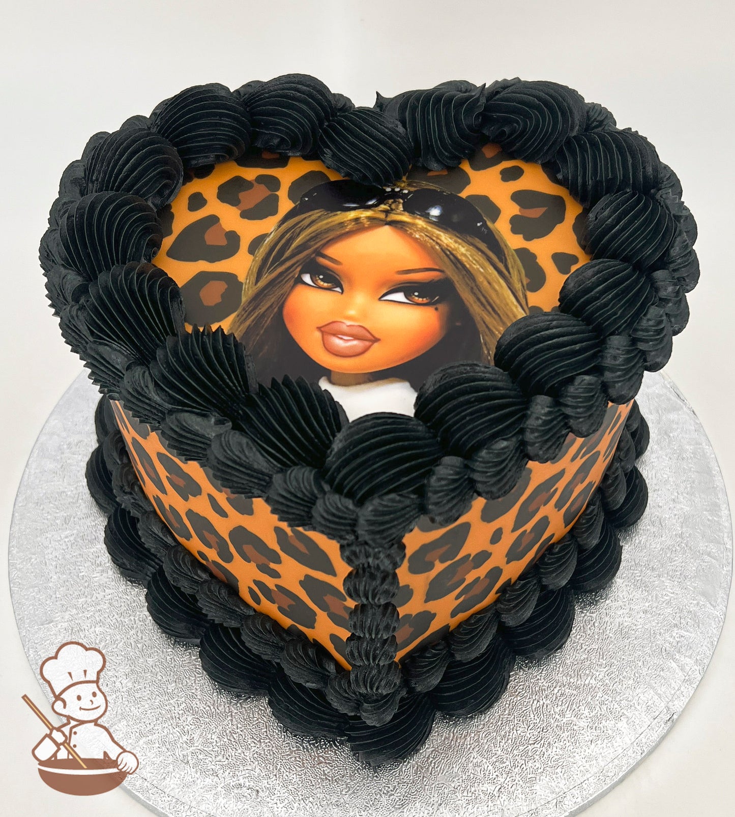 Heart-shaped cake with leopard print design, topped with a cartoon character image. Black frosting borders the top and sides, on a silver cake board.