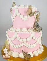 Two-tier pink cake with white piped frosting in scalloped and rosette designs, adorned with gold decorative butterfly toppers, on a gold cake board.