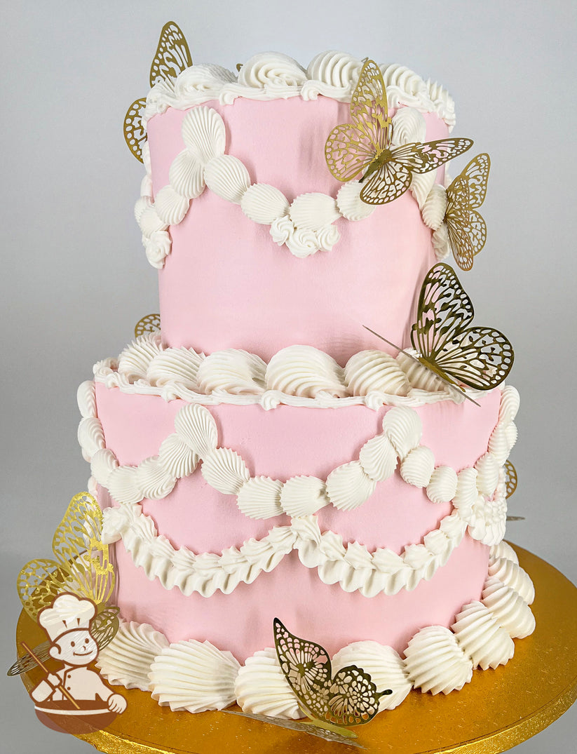 Two-tier pink cake with white piped frosting in scalloped and rosette designs, adorned with gold decorative butterfly toppers, on a gold cake board.