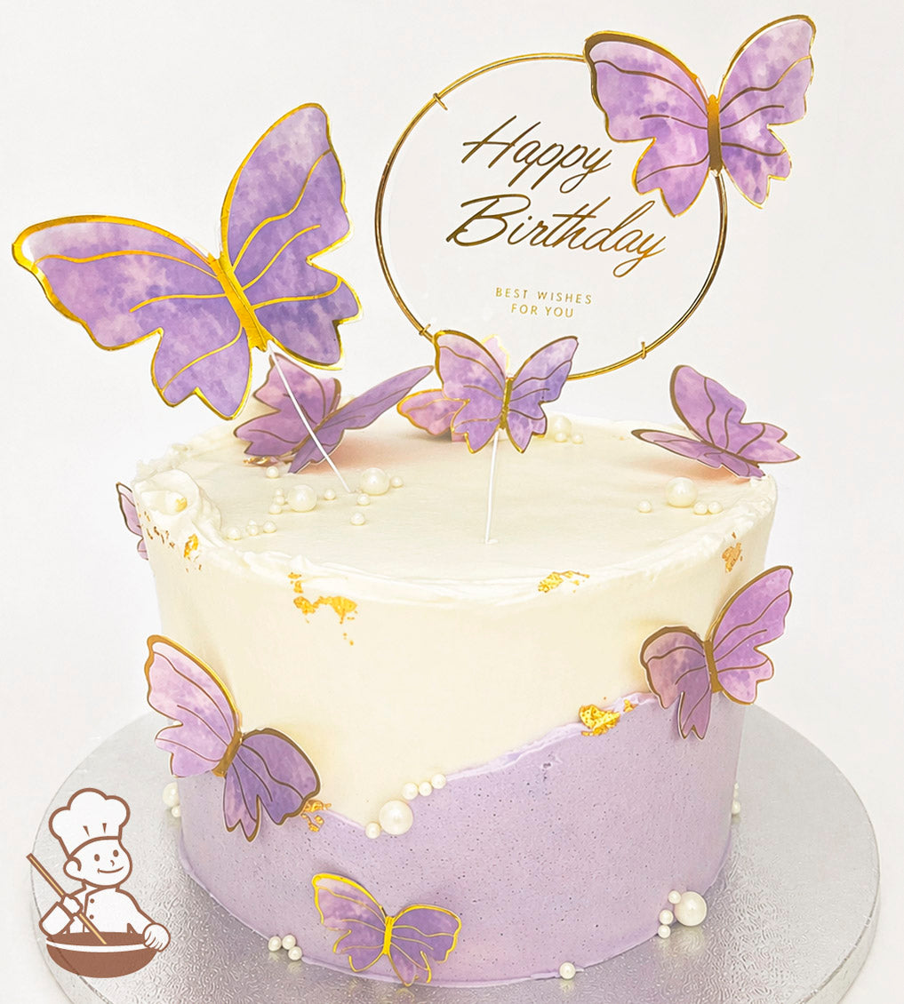 Round cake with white and lavender frosting, adorned with purple butterflies and pearl decorations. A "Happy Birthday" topper is on top.