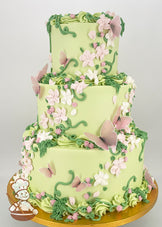 Three-tiered cake with light green frosting, adorned with pink and white buttercream flowers, green vines, and delicate pink butterfly decorations on a gold base.