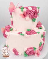 Two-tier pink cake decorated with pink and white buttercream swirls, green leaves, edible pearls, and delicate pink butterfly toppers.