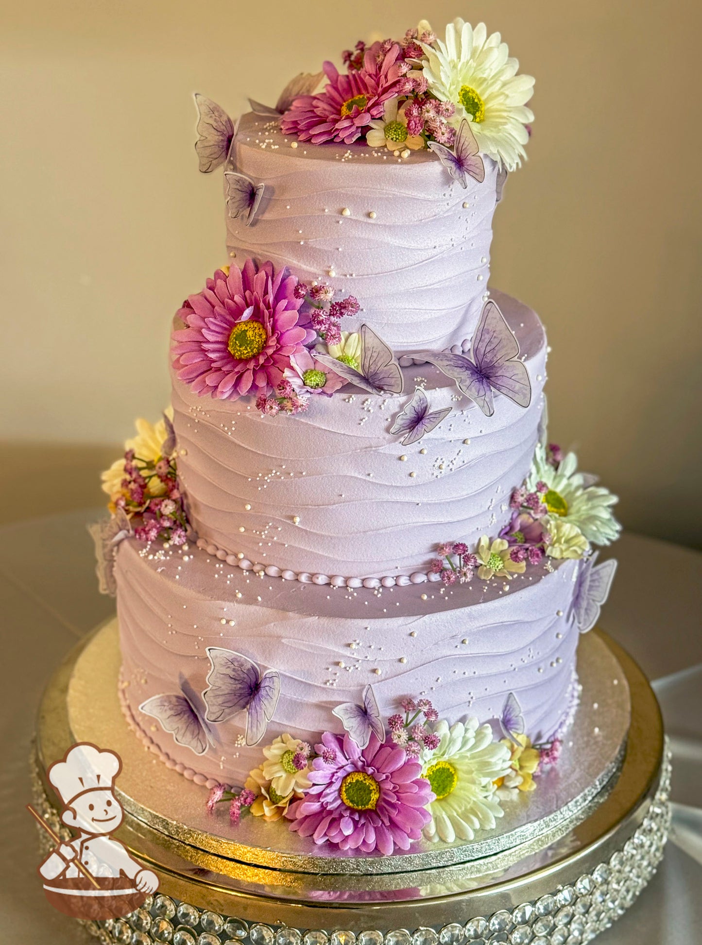 Three-tiered lavender cake with horizontal textured frosting, adorned with pink and white silk daisies, small silk flowers, edible pearls, and delicate purple butterfly decorations.