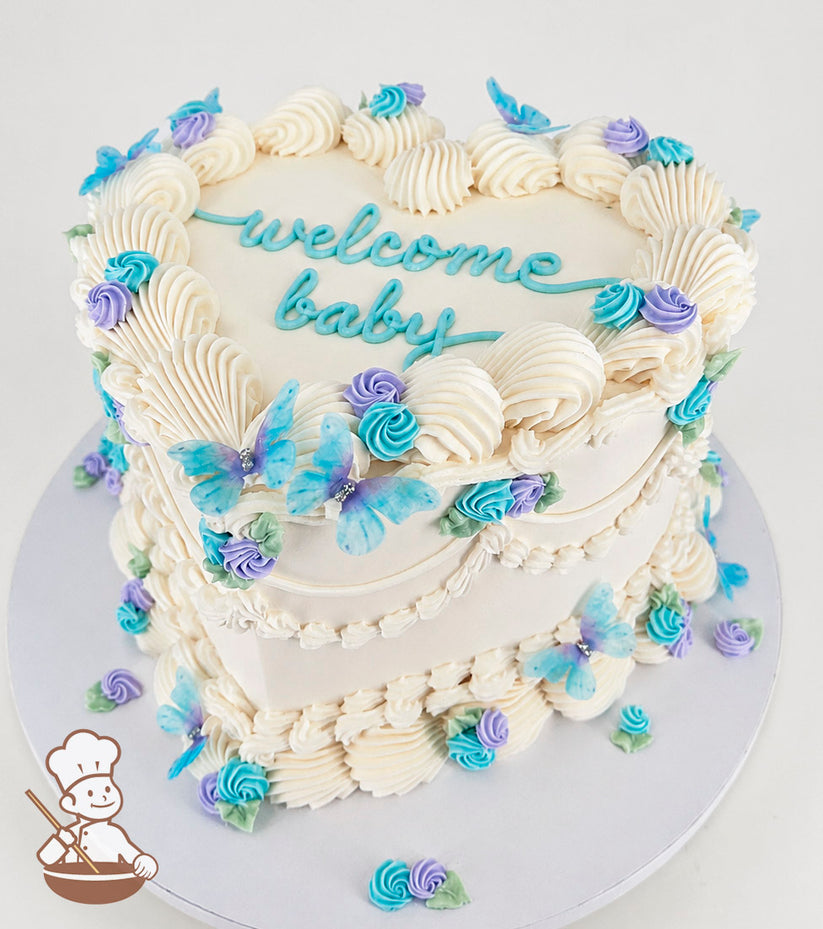 Heart-shaped, single tier white cake with piped swirls, baby blue and purple buttercream swirlettes, blue butterflies, and "welcome baby" written on top.