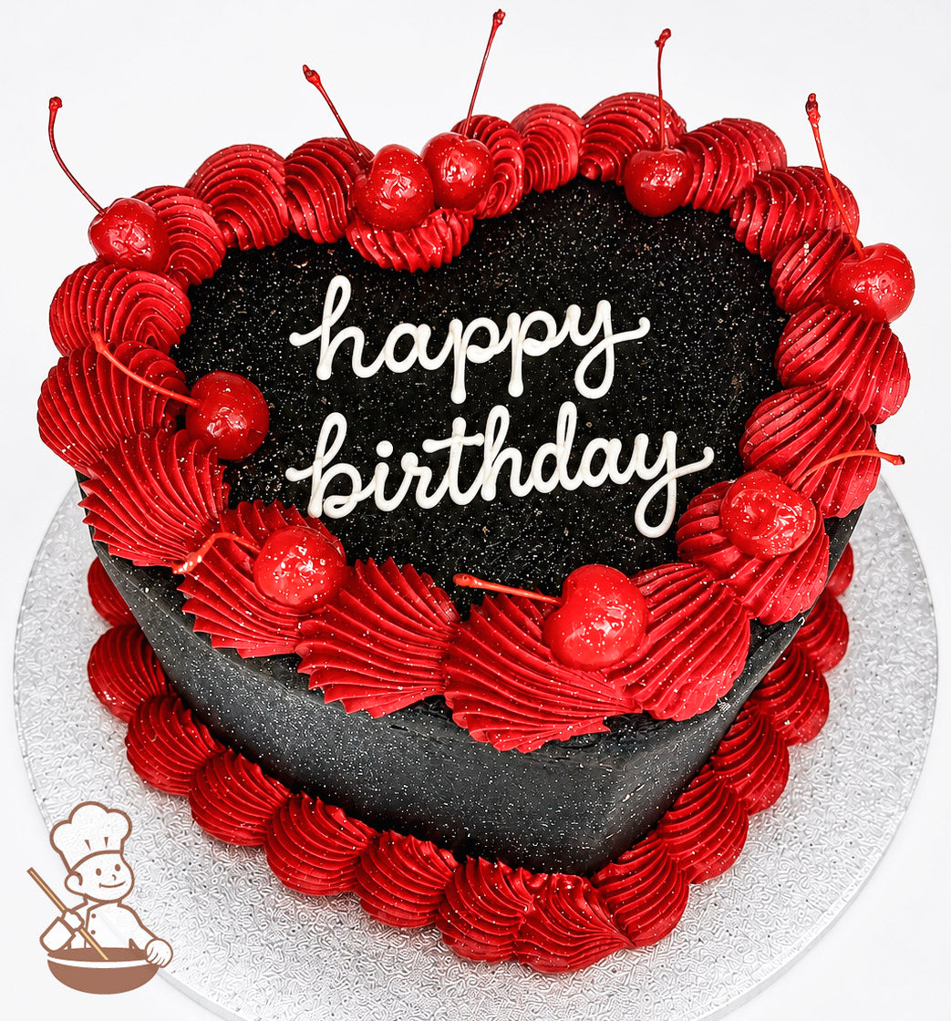 A heart-shaped black cake with "happy birthday" written in white icing on top. Decorated with red frosting swirls and cherries, placed on a silver textured board.