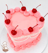 Heart-shaped pink cake with piped frosting swirls, topped with glossy red cherries. Decorated with pearl-like beads and set on a silver base.