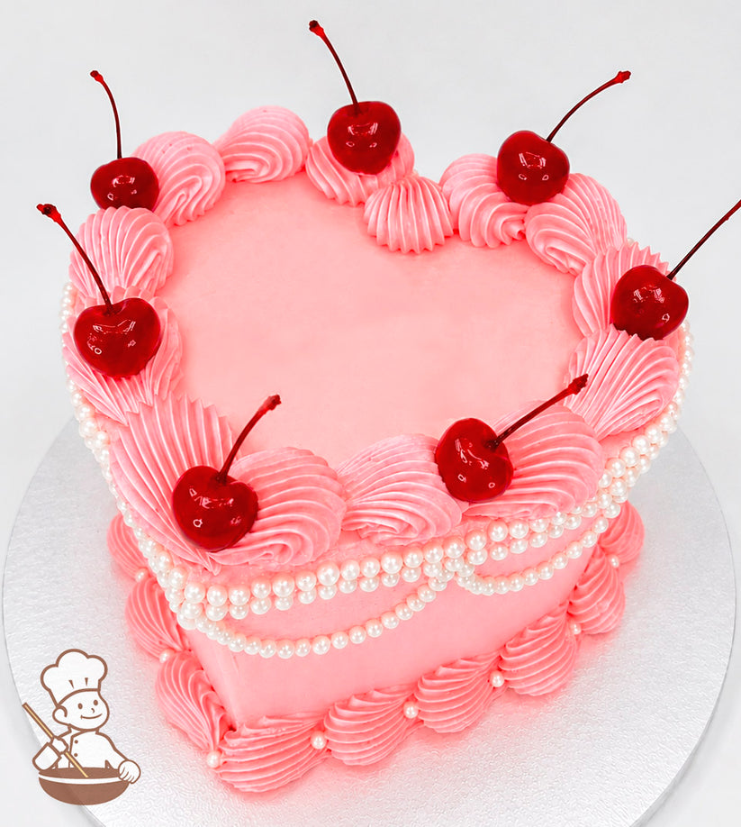 Heart-shaped pink cake with piped frosting swirls, topped with glossy red cherries. Decorated with pearl-like beads and set on a silver base.