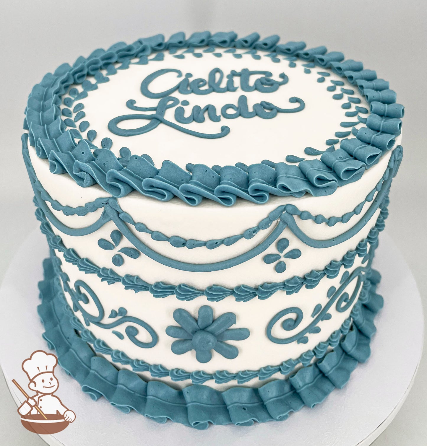 A single-tier white cake decorated with intricate blue buttercream piping, including floral patterns, swirls, and ruffled borders. The top features the text "Cielito Lindo" in blue icing.