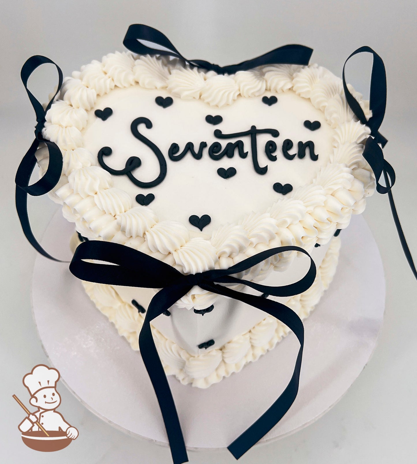 Heart-shaped cake with white frosting, black "Seventeen" text, small black hearts, and black ribbon bows. Decorated with piped frosting edges.