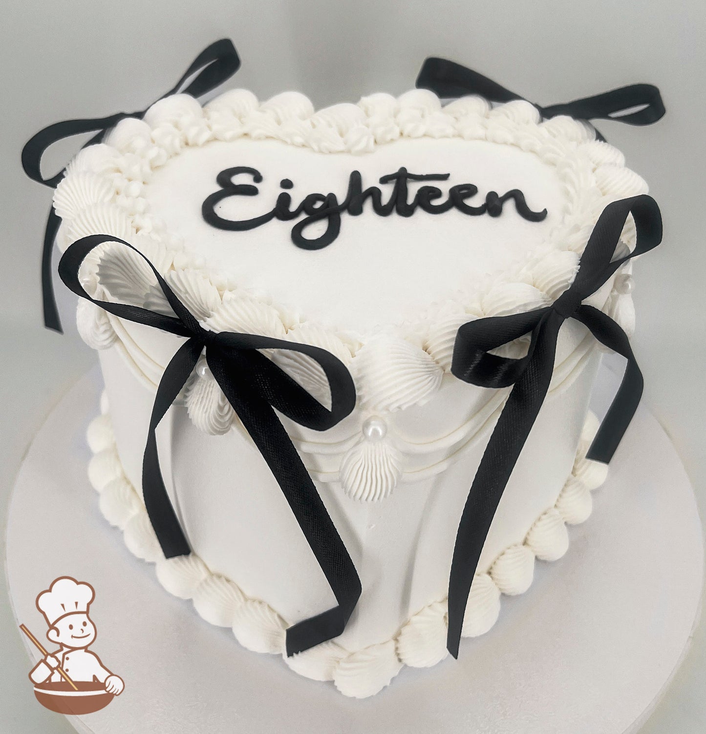 Heart-shaped white cake with "Eighteen" written on top, decorated with black ribbons and piped frosting borders.