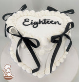 Heart-shaped white cake with "Eighteen" written on top, decorated with black ribbons and piped frosting borders.