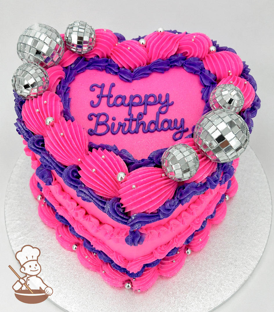 A heart-shaped, single-tier cake with bright pink and purple frosting, silver disco ball decorations, and "Happy Birthday" written in purple icing on top.