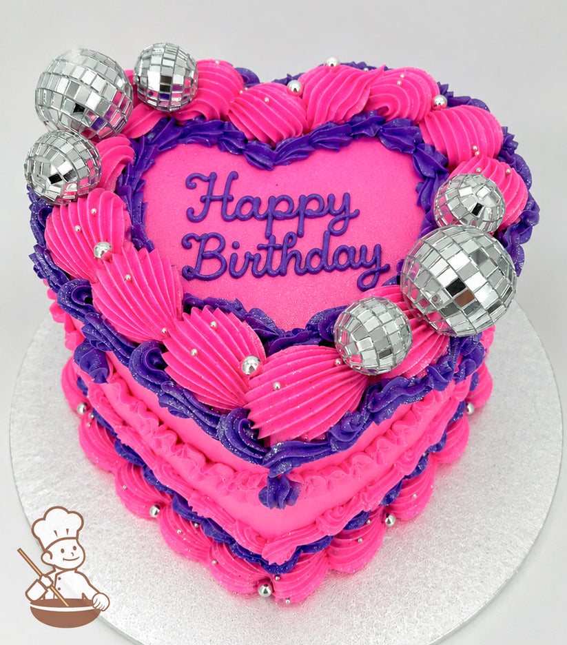A heart-shaped, single-tier cake with bright pink and purple frosting, silver disco ball decorations, and "Happy Birthday" written in purple icing on top.