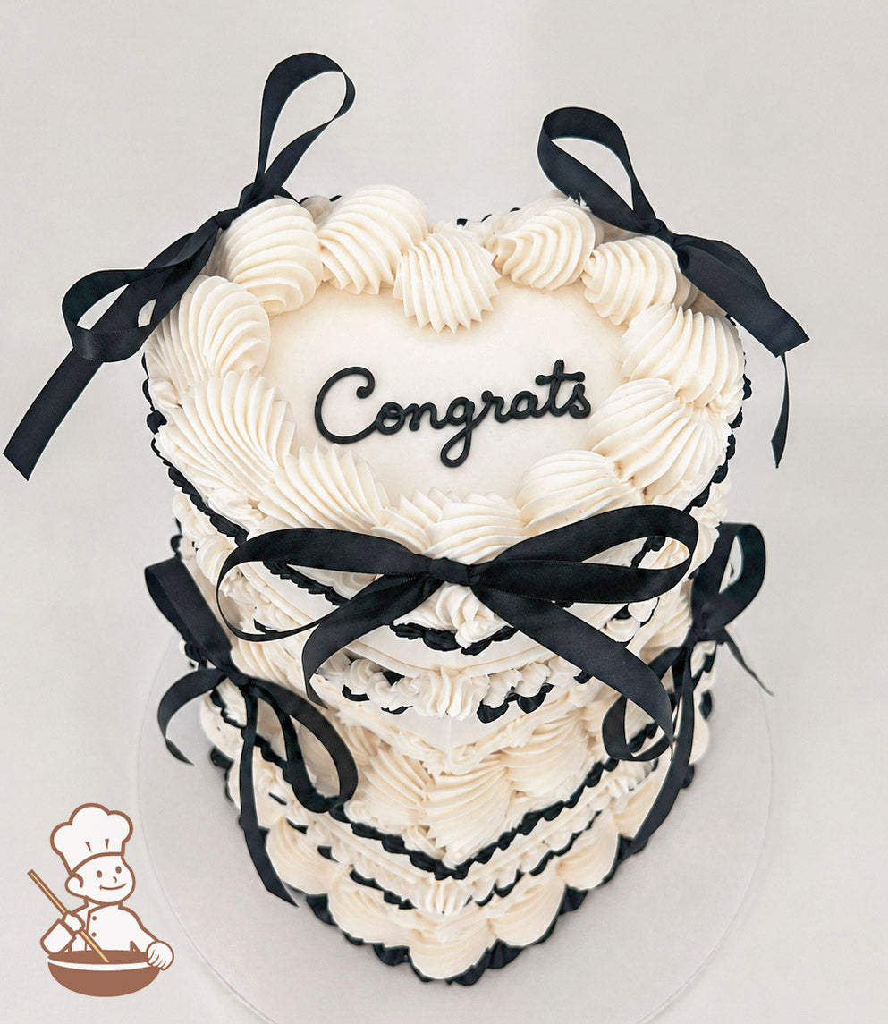 A two-tiered heart-shaped cake with white frosting, black piped accents, and black ribbon bows. The top tier features the word "Congrats" in black script.