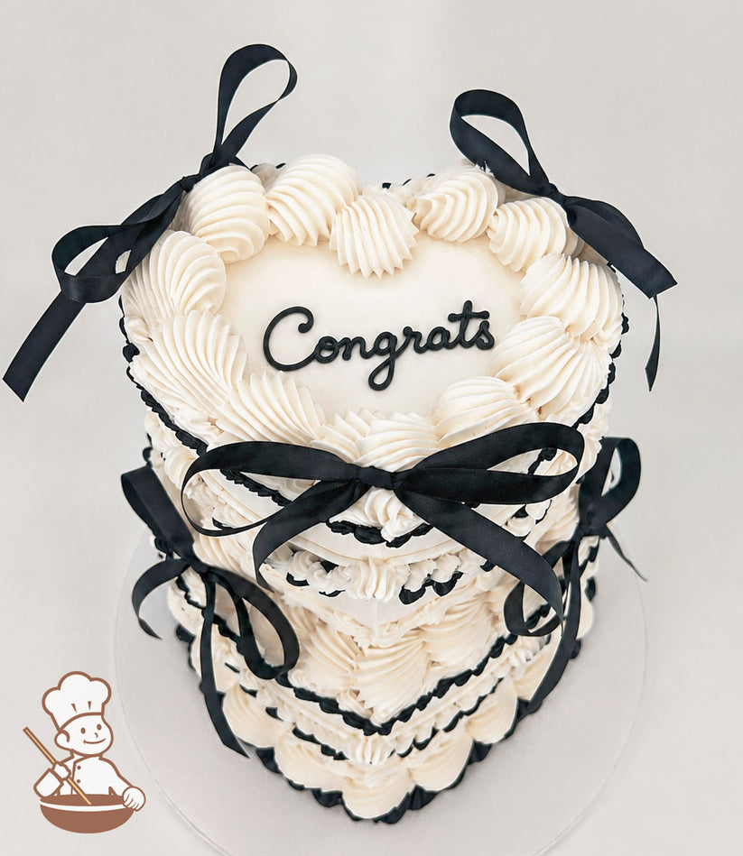 A two-tiered heart-shaped cake with white frosting, black piped accents, and black ribbon bows. The top tier features the word "Congrats" in black script.