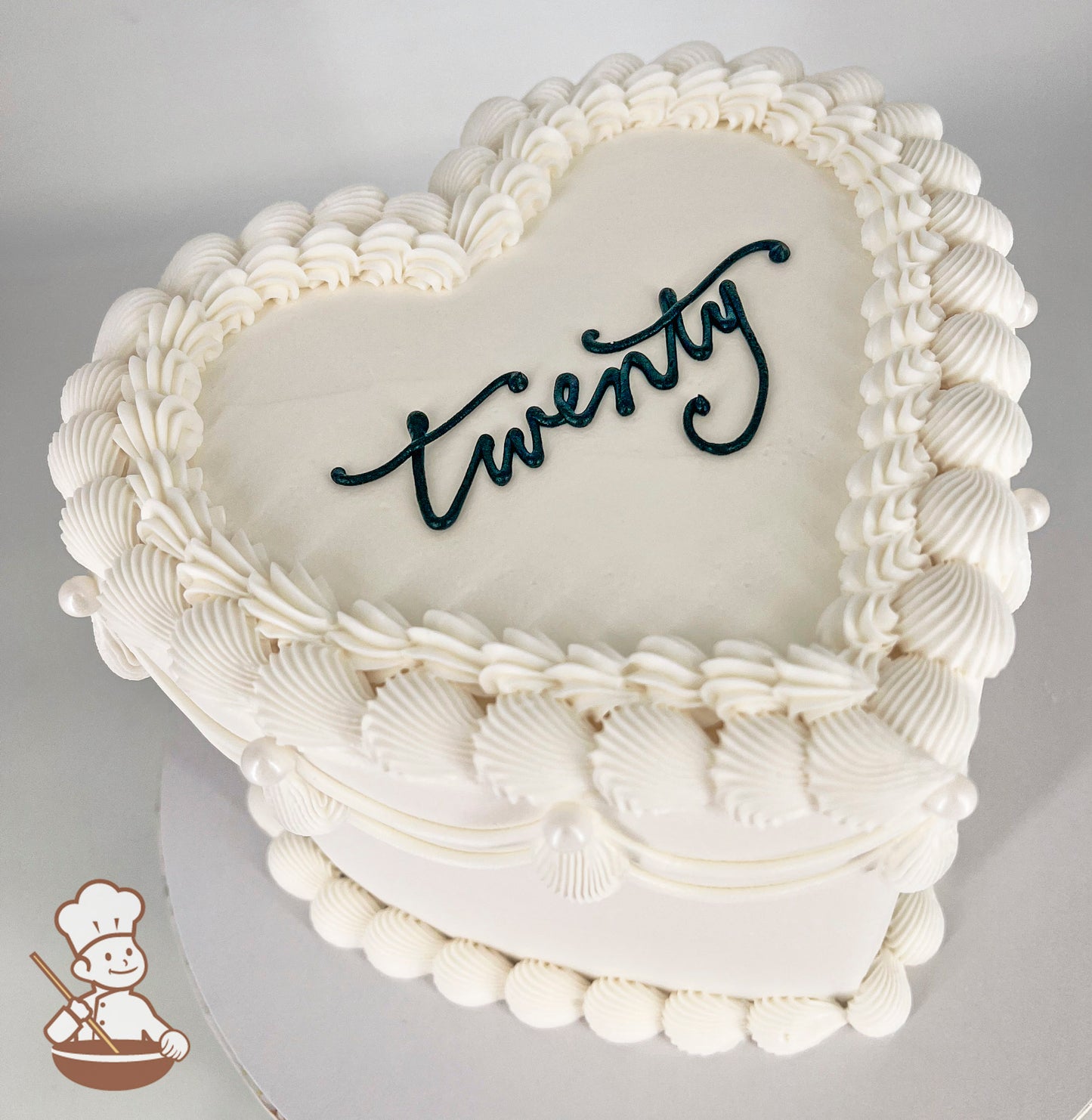 Heart-shaped white cake with piped shell and pearl-like frosting details. The word "twenty" is written in elegant black script on top.