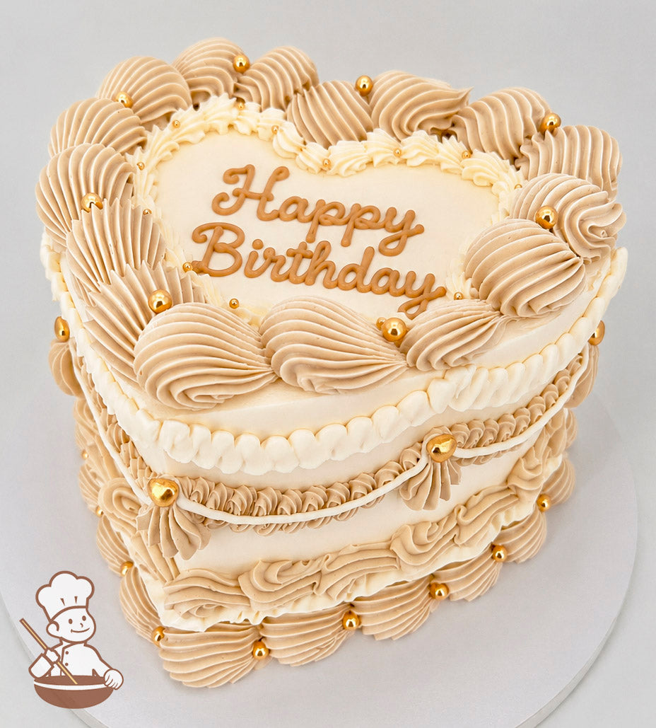 Heart-shaped cake with beige frosting swirls, gold beads, and "Happy Birthday" text on top. Placed on a white cake board.