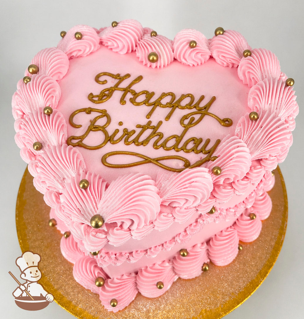 Heart-shaped pink cake with "Happy Birthday" written in gold icing. Decorated with pink piped frosting swirls and gold pearl accents on a gold cake board.
