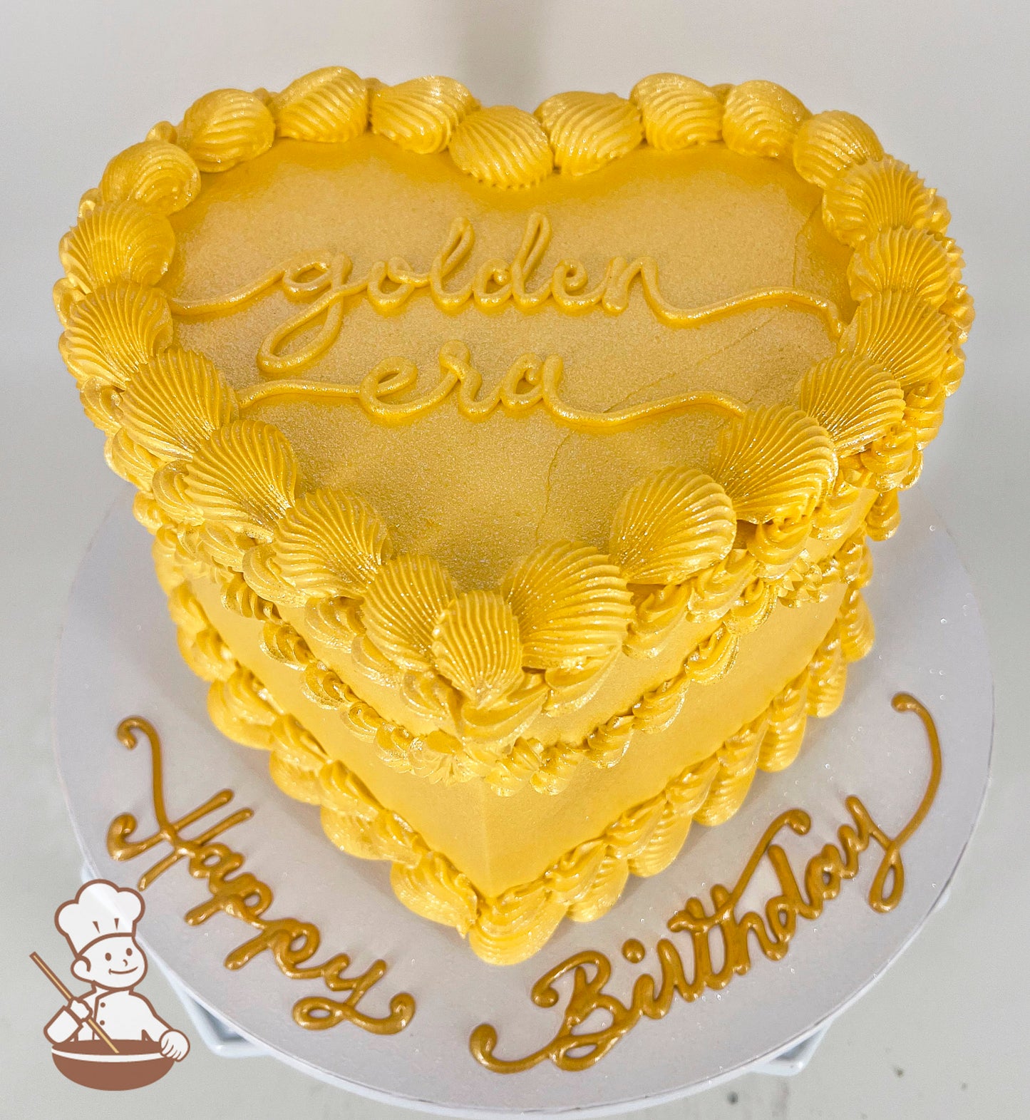 A heart-shaped, single-tiered golden cake with shell-like frosting details. "Golden Era" is written on top, and "Happy Birthday" decorates the base.