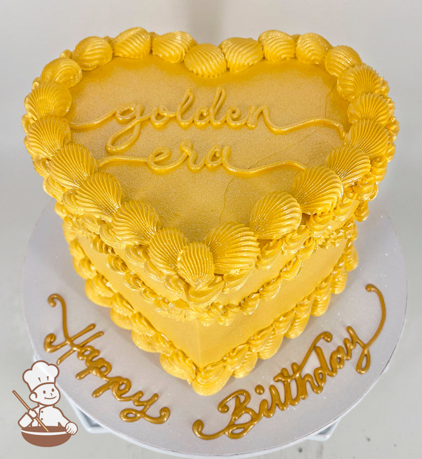 A heart-shaped, single-tiered golden cake with shell-like frosting details. "Golden Era" is written on top, and "Happy Birthday" decorates the base.