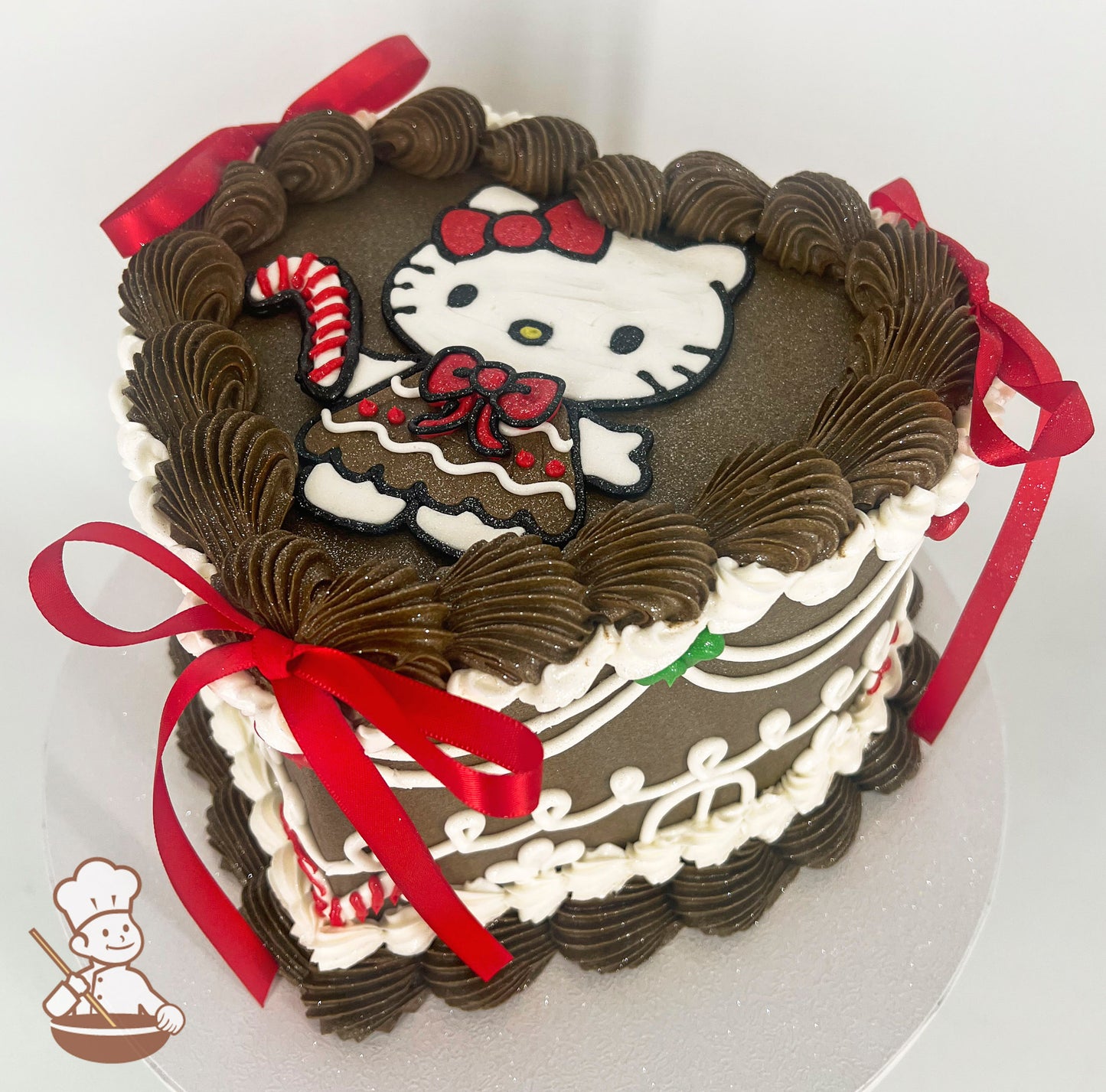 Heart-shaped brown cake with piped brown and white frosting, featuring a Hello Kitty design on top. Decorated with red satin bows and candy cane accents.