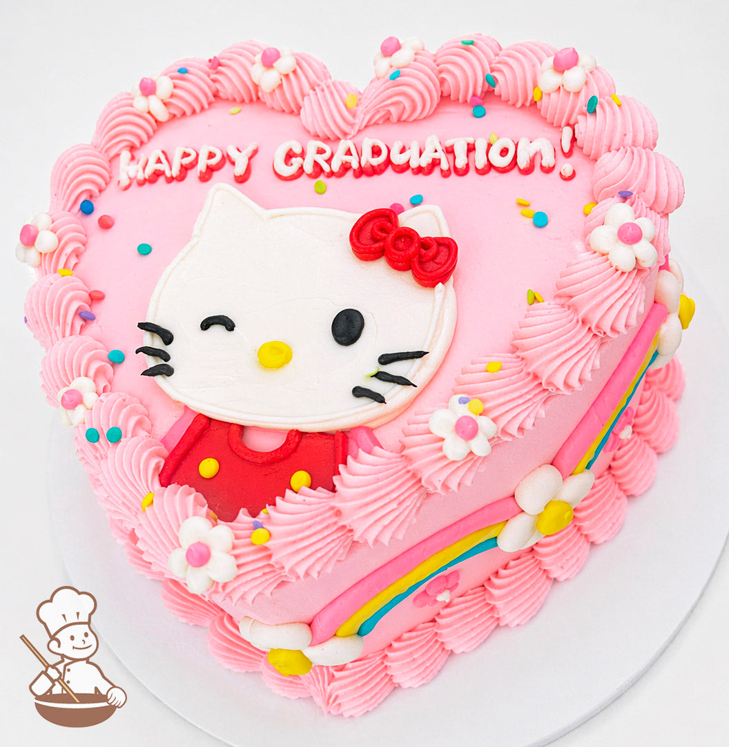 Heart-shaped pink cake with "Happy Graduation!" text, featuring a winking Hello Kitty design, piped pink frosting, rainbow accents, and small flower decorations.