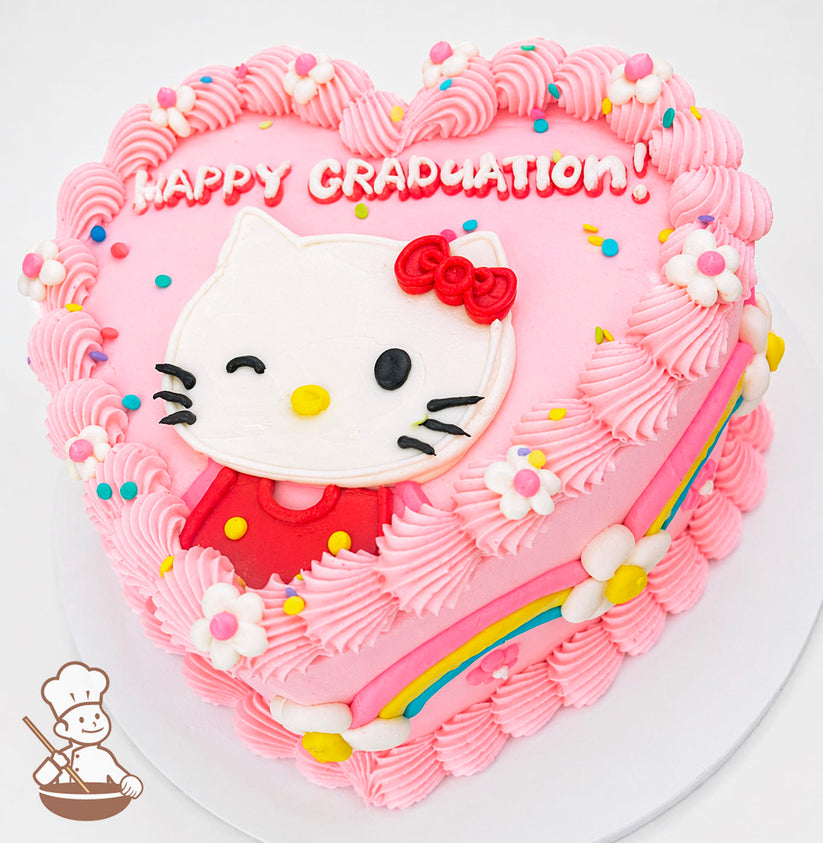 Heart-shaped pink cake with "Happy Graduation!" text, featuring a winking Hello Kitty design, piped pink frosting, rainbow accents, and small flower decorations.