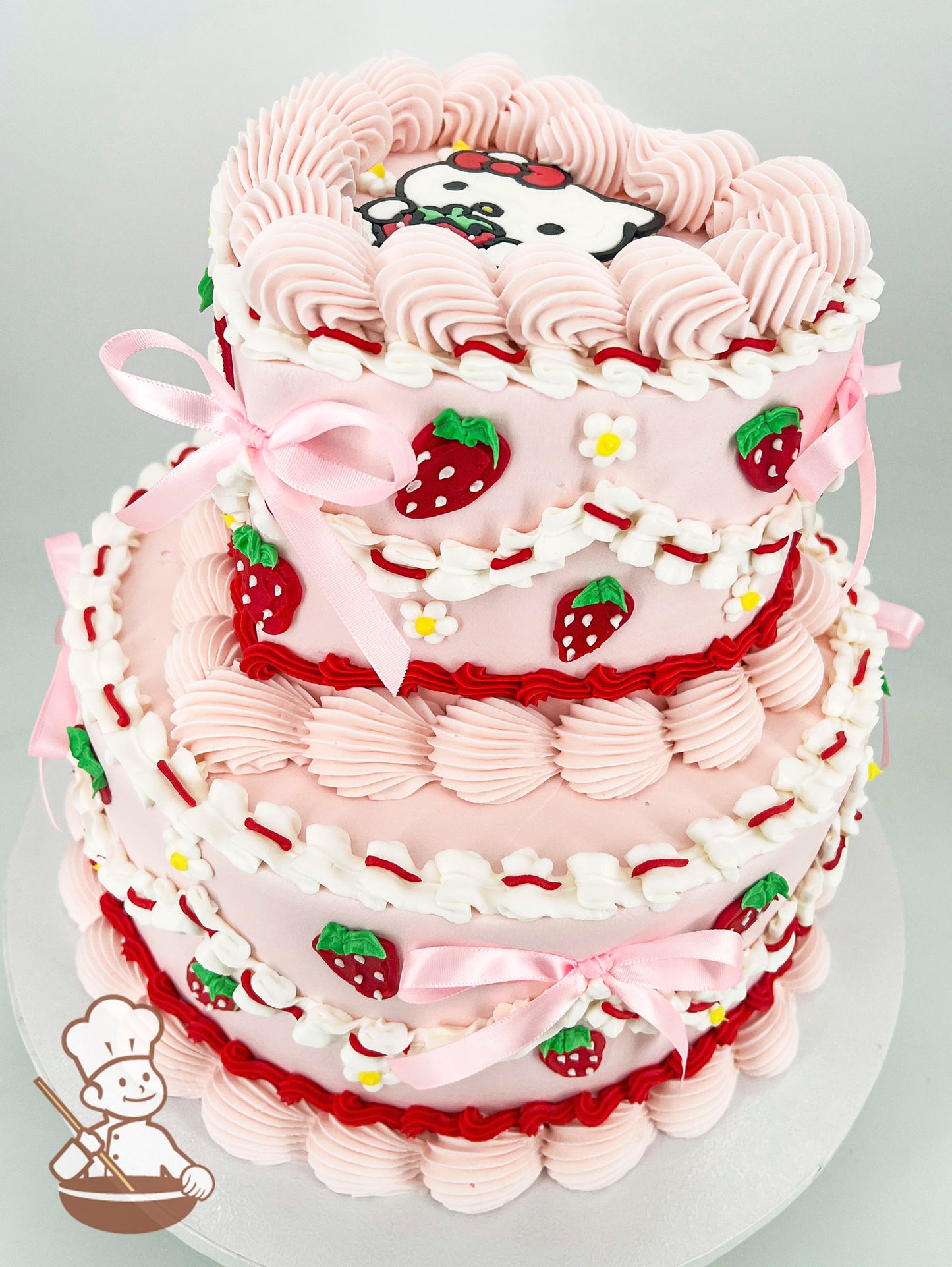 A two-tier pink cake decorated with piped strawberries, small flowers, pink ribbons, and a cartoon character on the top tier.