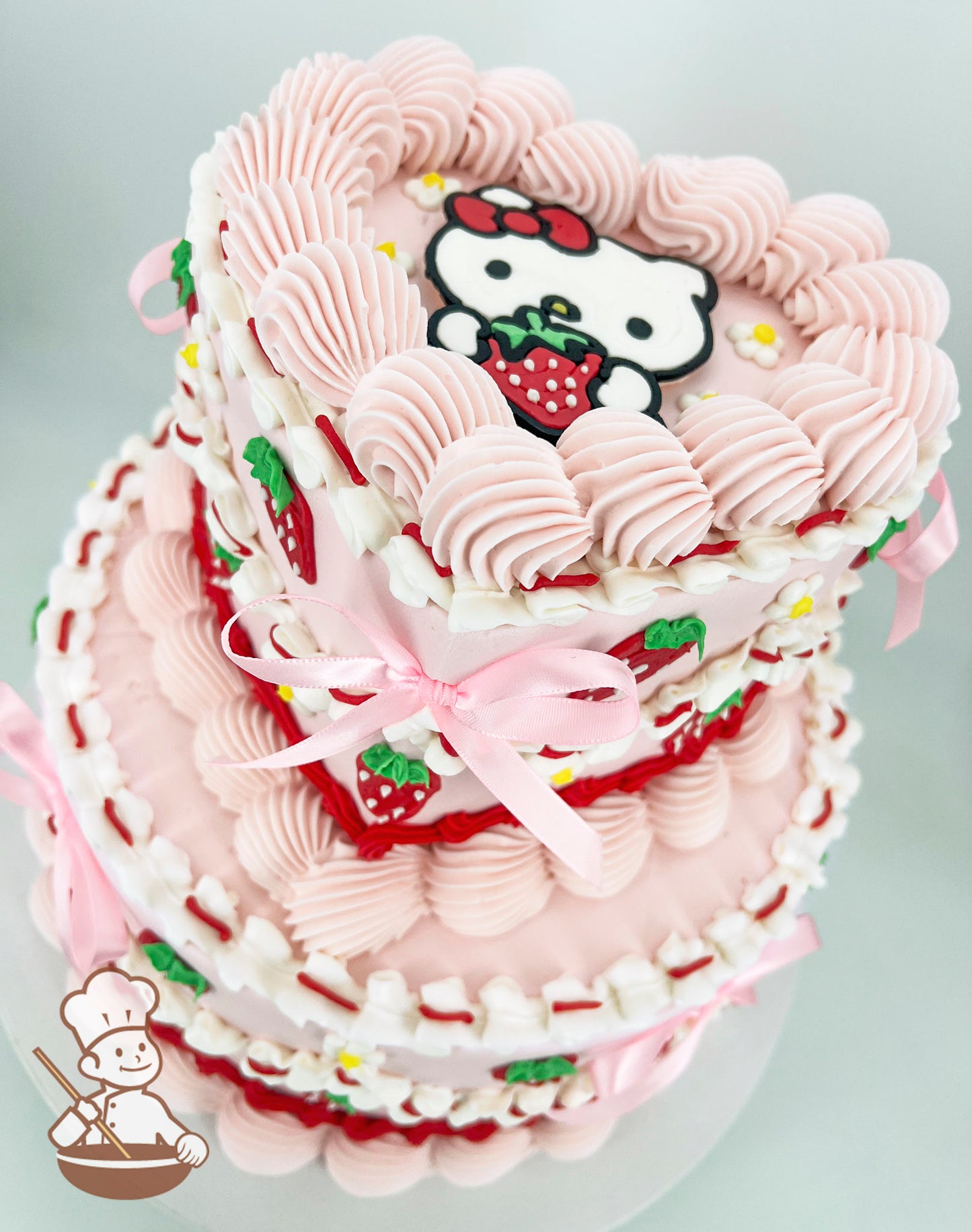 A two-tier heart and round shaped cake with pink frosting, piped swirls, strawberry decorations, and a Hello Kitty topper holding a strawberry. Pink ribbons accent the design.