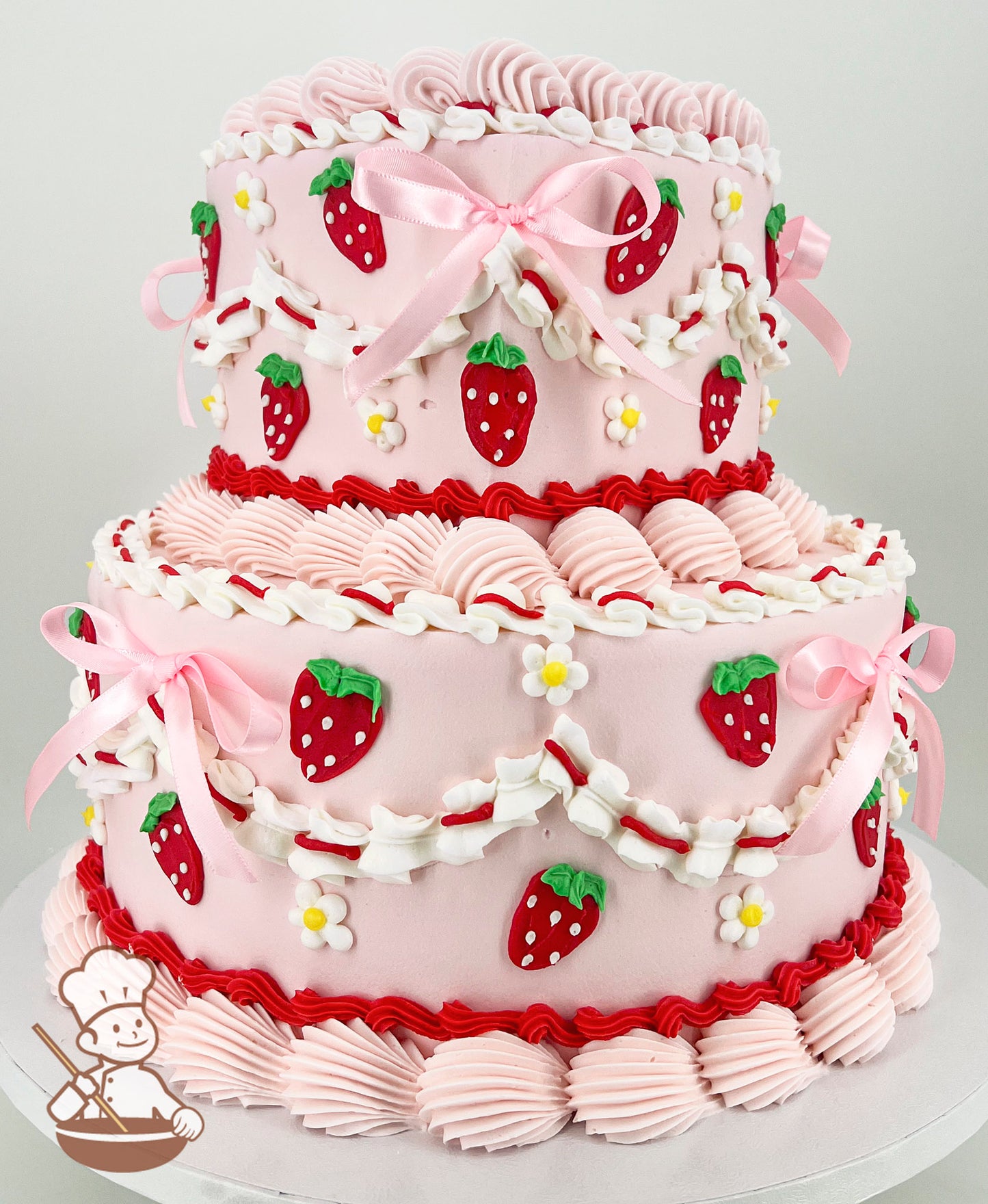 Two-tier pink cake with strawberry decorations, white and red icing swirls, pink bows, and small white flowers on a white board.