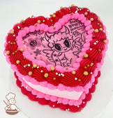 Heart-shaped cake with pink and red piped frosting, adorned with gold and white sprinkles. The top features a cartoon-style illustration of a winged character.