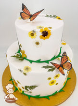 Two-tier white cake adorned with yellow and white flowers, green leaf accents, and realistic orange butterfly toppers. Gold cake board.