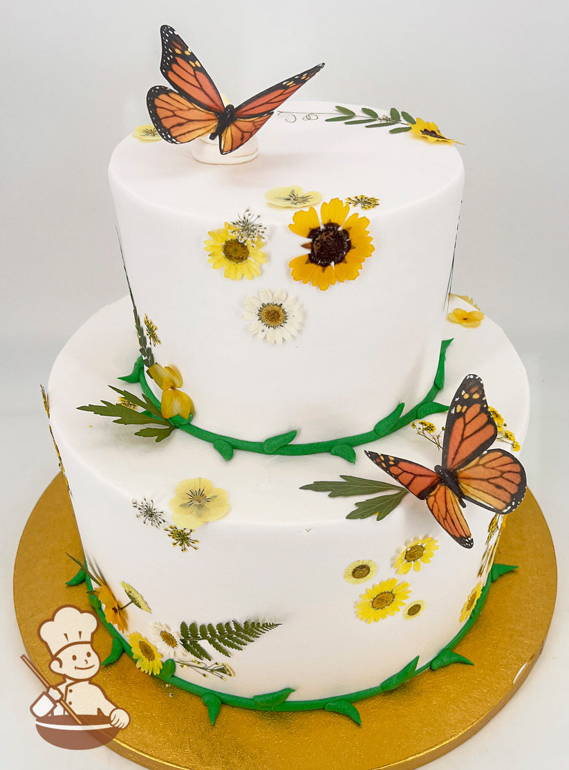 Two-tier white cake adorned with yellow and white flowers, green leaf accents, and realistic orange butterfly toppers. Gold cake board.