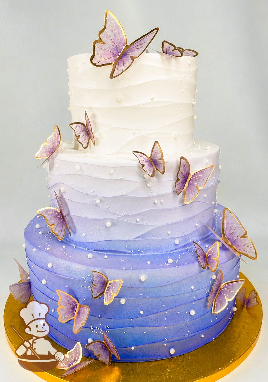 A three-tier cake with a gradient design transitioning from white to purple. Decorated with purple butterflies with gold edges and pearl accents.