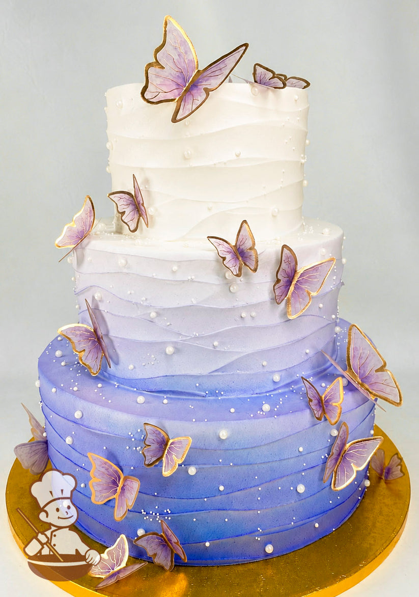 A three-tier cake with a gradient design transitioning from white to purple. Decorated with purple butterflies with gold edges and pearl accents.