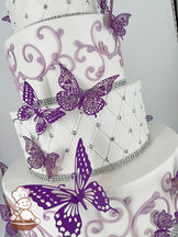 A multi-tier white cake with intricate lilac scrollwork, silver bead accents, and vibrant purple butterfly decorations.