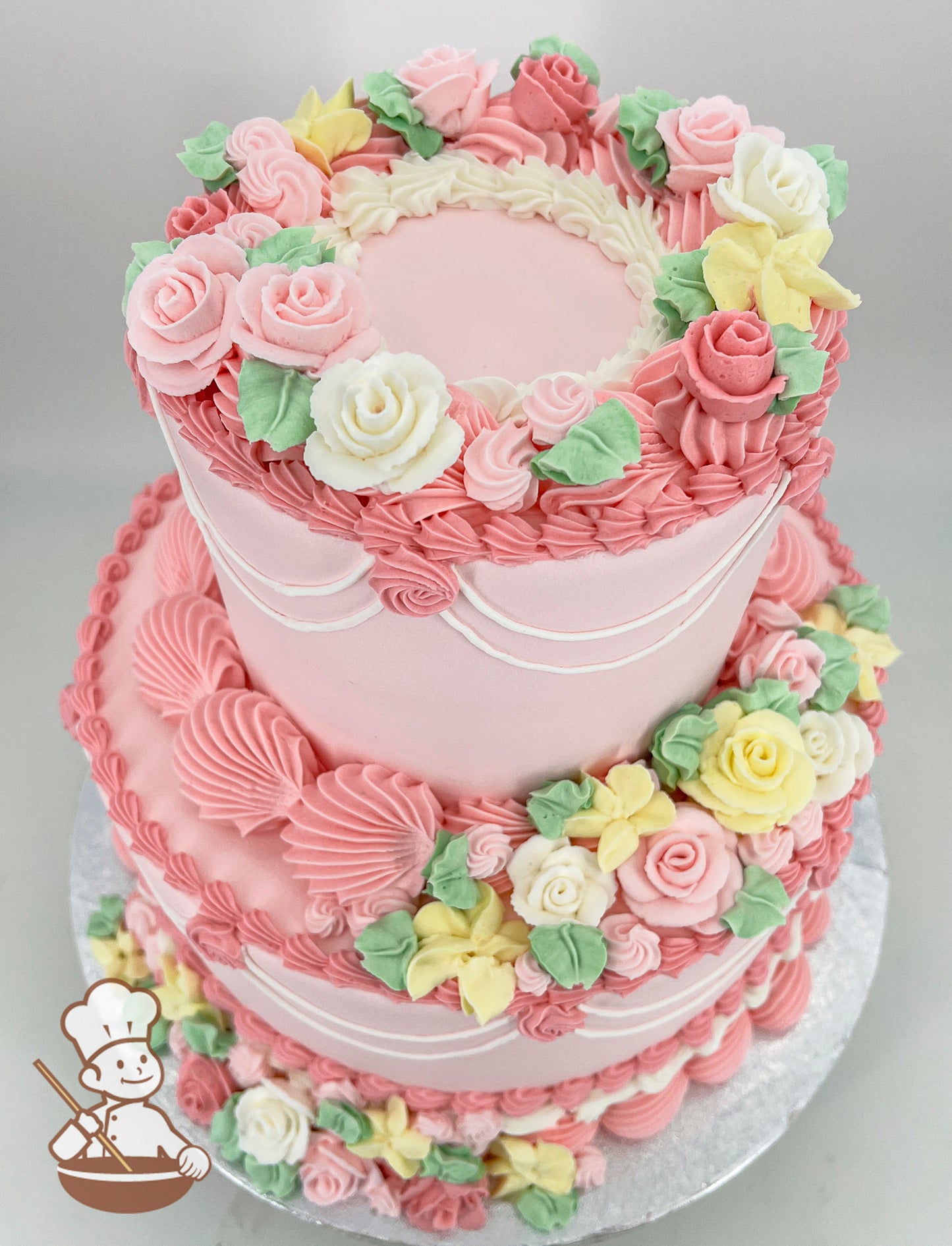 Two-tier pink cake decorated with intricate buttercream flowers in pastel pink, yellow, and white, with green leaves and piped details on a silver base.