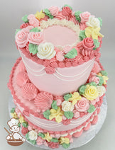 Two-tier pink cake decorated with intricate buttercream flowers in pastel pink, yellow, and white, with green leaves and piped details on a silver base.