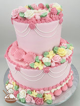 Two-tier pink cake decorated with piped buttercream roses, leaves, and swirls in pastel pink, yellow, white, and green, with white icing drapes.