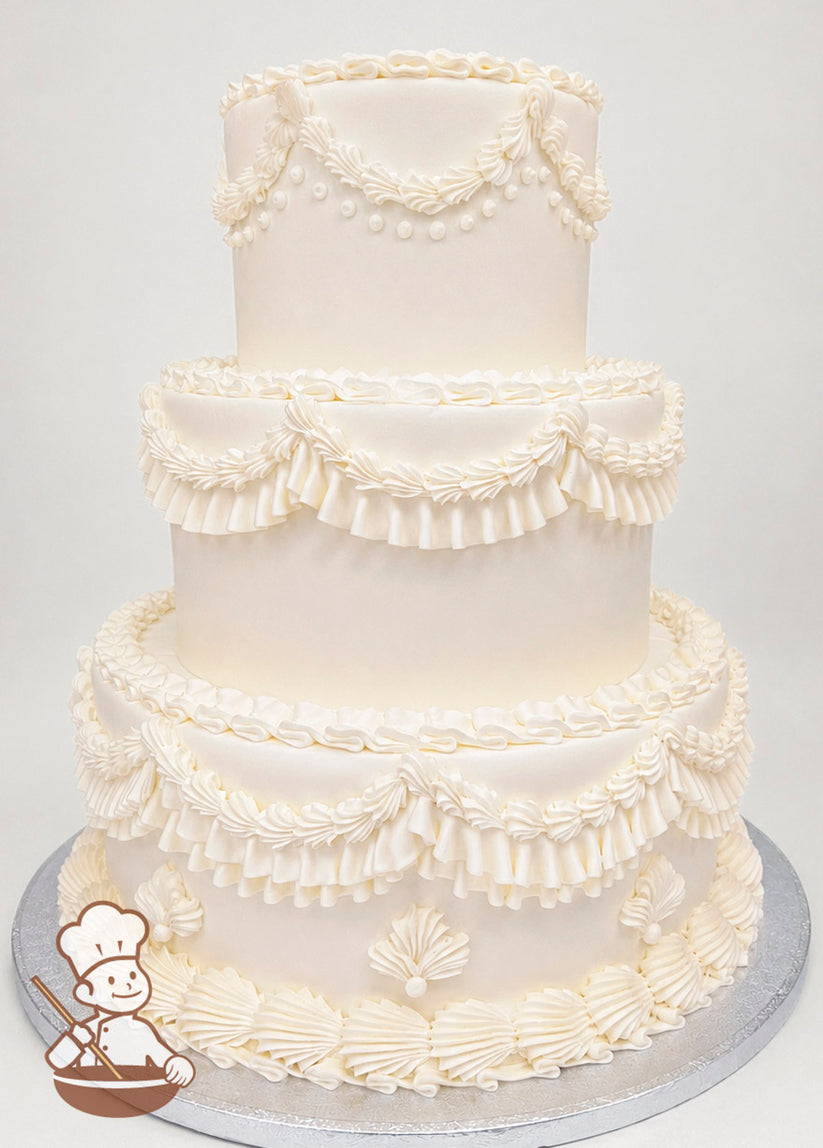Three-tier white cake with intricate piped buttercream details, including ruffled swags, pearls, and floral patterns, on a silver base.