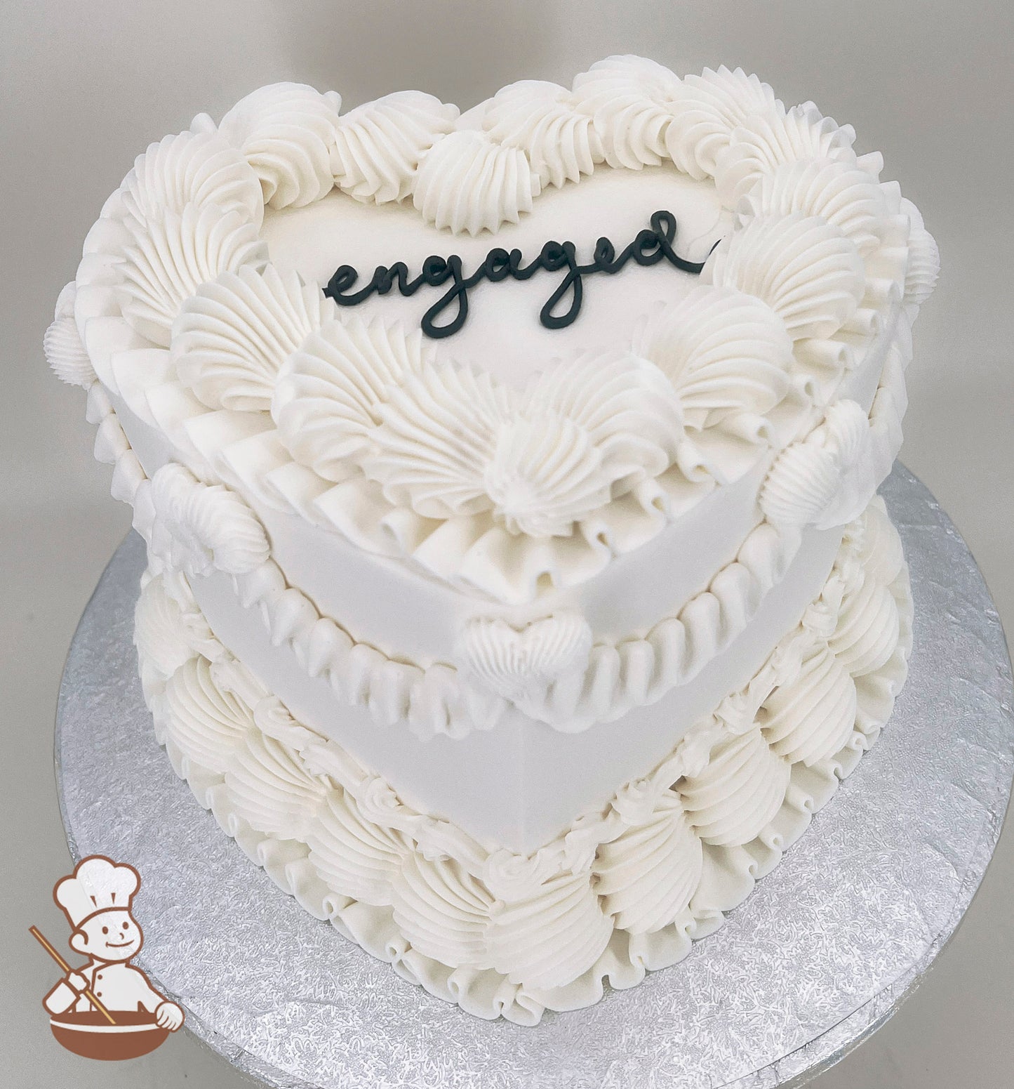 A heart-shaped, single-tier white cake with intricate piped frosting details and the word "engaged" written in black icing on top.